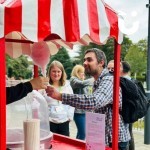 Candy Floss Cart Hire 1 set up outside at a corporate event with a fun expert hading a stick of candy floss over to a fun seeker